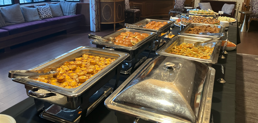 A close up photo of the buffet table, with many silver dishes lining two sides of a long rectangular table. Snacks include cheese pastry cups, flatbread, and sliders.