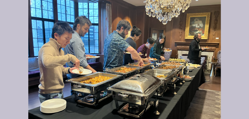 A photo of six people going down the buffet line, taking small bites from silver containers and adding them to their paper plates