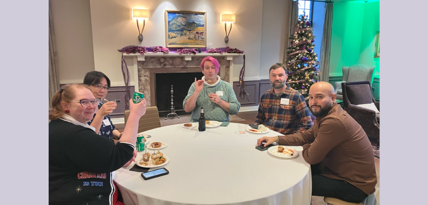A group of people from Life Sciences sits around a white table with plates of food. They smile for a camera with a fireplace and tree in the background.